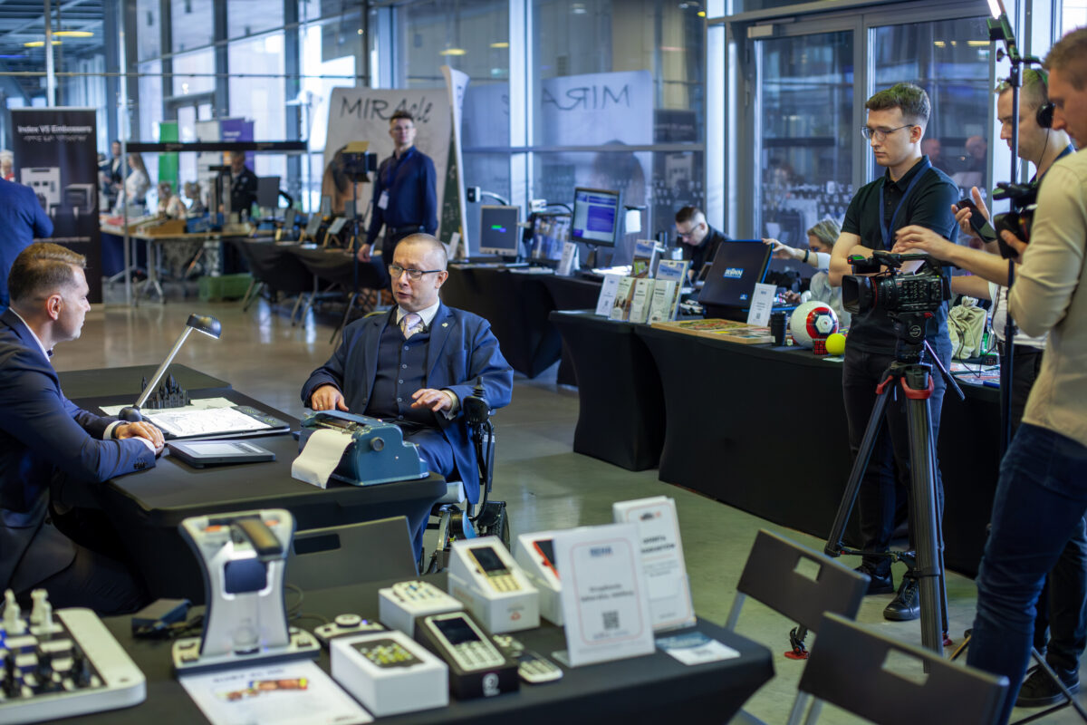 Hala Accessibility Expo 2025: stoiska z urządzeniami brajlowskimi i elektroniką, przy stole wystawca, w tle ekipa filmowa z kamerą.