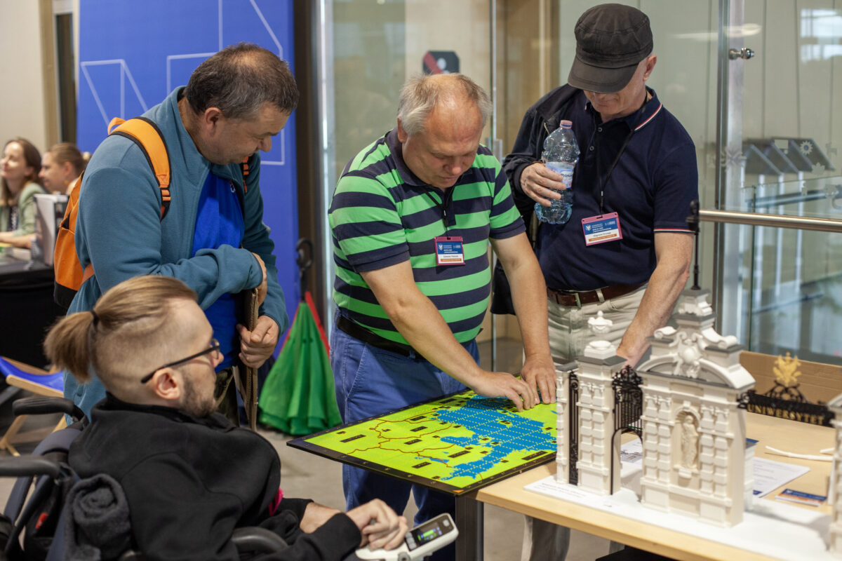 Zwiedzający, w tym mężczyzna na wózku, dotykają wypukłej mapy i białej makiety budynku na stoisku Accessibility Expo 2025.