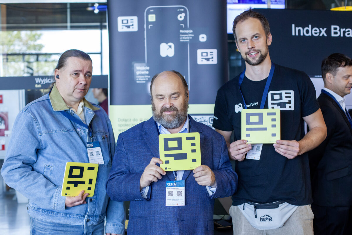 Trzej mężczyźni pokazują żółte tabliczki z dotykowymi kodami przy stoisku Index Braille na Accessibility Expo 2025.
