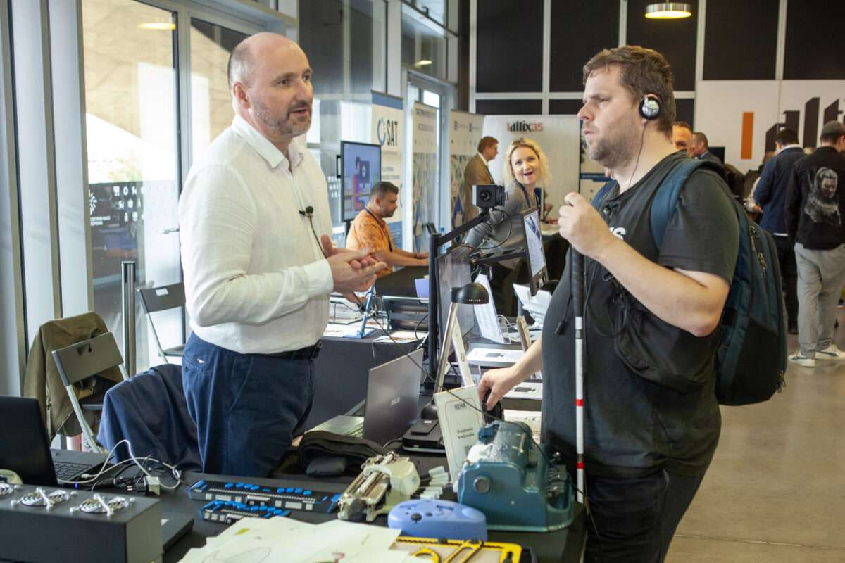 Wystawca prezentuje urządzenie zwiedzającemu z białą laską przy stoisku z elektroniką brajlowską na Accessibility Expo 2025.