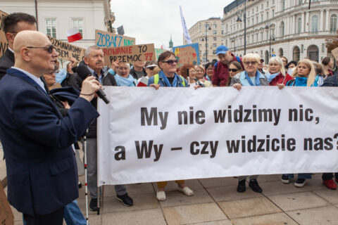 Uczestnicy manifestacji z transparentem „My nie widzimy nic, a Wy – czy widzicie nas?”; na pierwszym planie mężczyzna z mikrofonem i białą laską.
