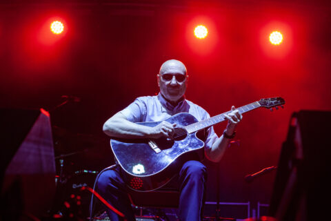 Gitarzysta w ciemnych okularach gra na gitarze akustycznej na scenie w czerwono-niebieskim świetle podczas koncertu Mazovia Stage 2025.