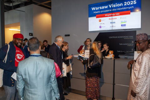 Uczestnicy przy stanowisku informacyjnym pod ekranem „Warsaw Vision 2025 – Miasto przyjazne dla niewidomych”.